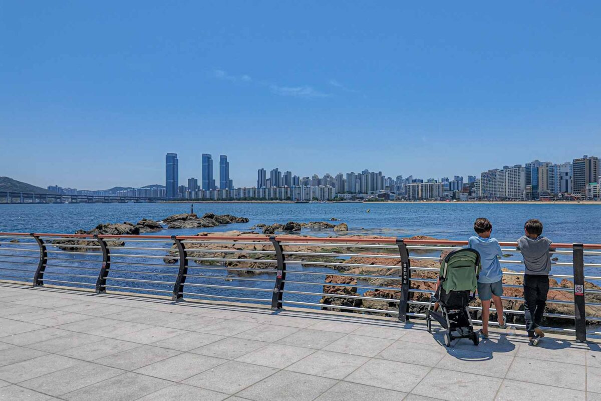 Two children stand beside a stroller at the waterfront railing in Gwangan Beach Park with the Busan skyline and bridge across the water. The broad paved path and open waterfront show one of the easier places for getting around Busan with kids on foot.