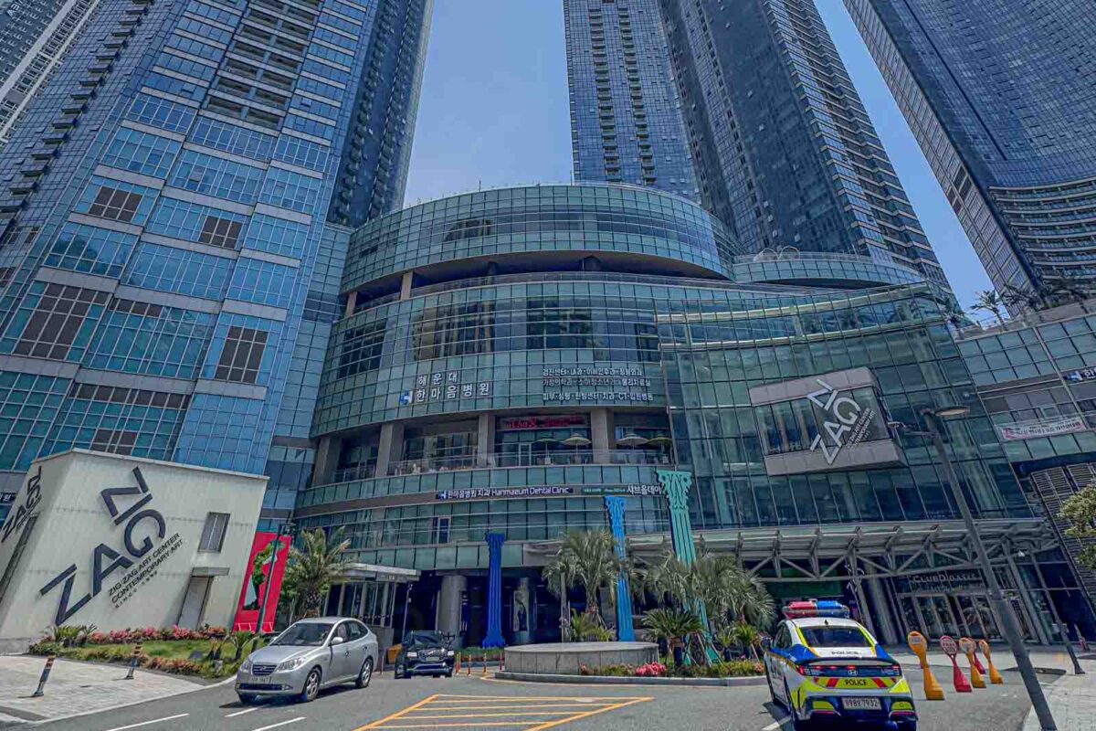 Tall glass LCT towers rise above a driveway and mall entrance in Haeundae with cars waiting below. Large complexes like this can have multiple entrances and pickup areas that families need to check carefully when arranging a taxi or Uber.