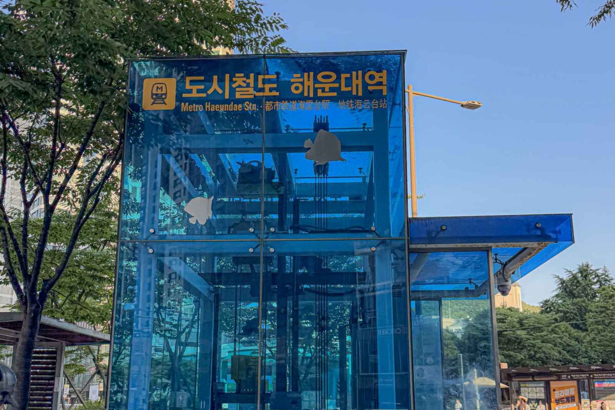 A blue glass elevator entrance above Haeundae Metro Station sits beneath leafy trees. The sign reads "도시철도 해운대역". "Metro Haeundae Stn". Street level elevators make the Busan Metro more manageable with strollers and luggage.