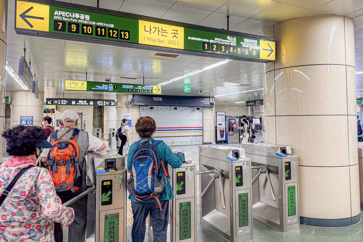 Passengers tap through a row of Busan Metro fare gates beneath large yellow and green directional signs. The signs read "APEC Naru Park". "Way Out". And show numbered exits including "1". "2". "3". "4". "5". "6". "8". "7". "9". "10". "11". "12". "13".