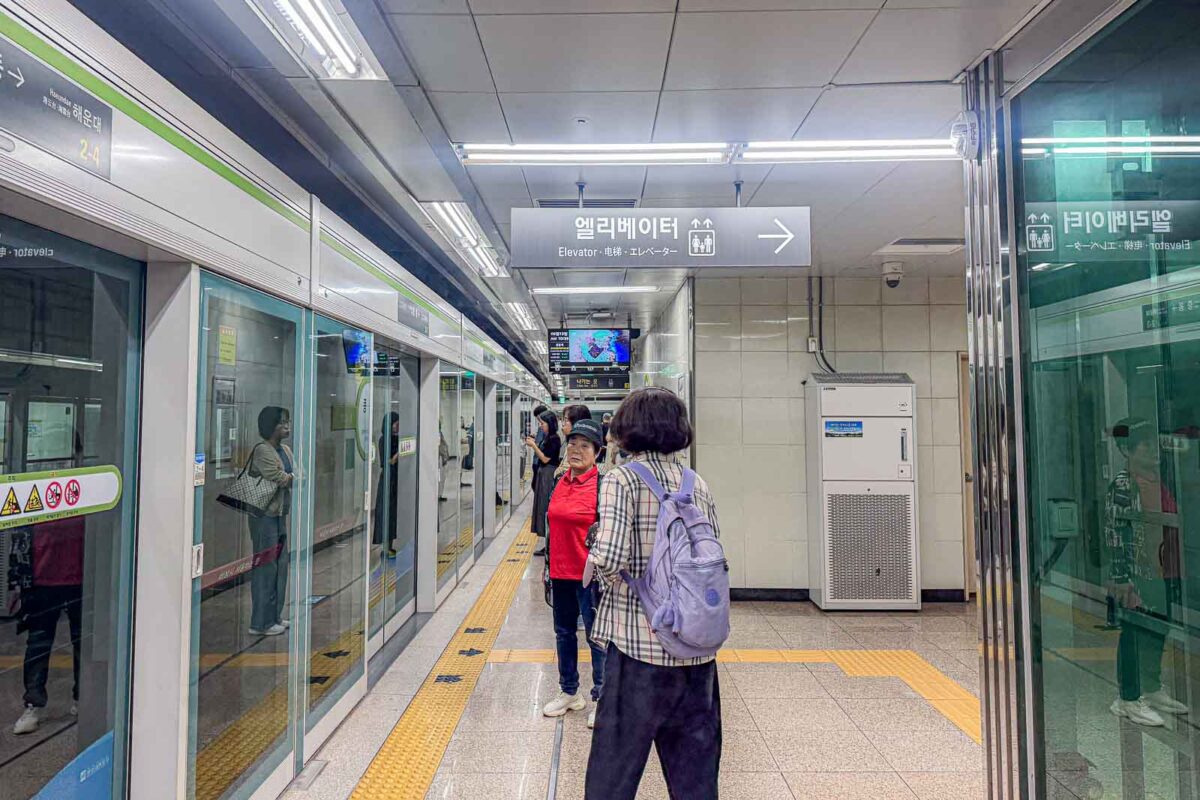 Passengers wait behind glass platform doors at Jungdong Station beside yellow tactile paving. An overhead sign reads "Elevator" with an arrow pointing toward the accessible route.