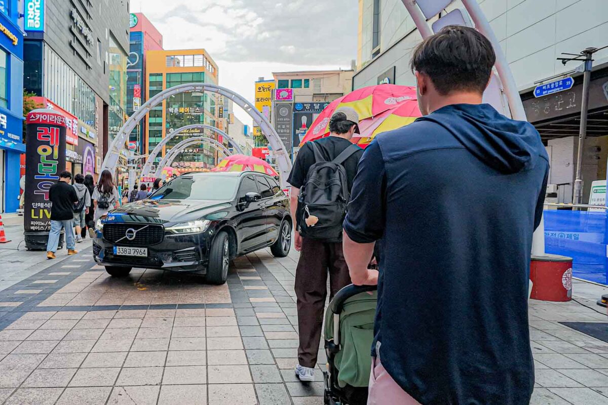 An adult pushes a stroller through a busy pedestrian area at BIFF Square while a car slowly passes through the same space. The crowded street shows why families need to stay aware of vehicles even in pedestrian focused parts of Busan.