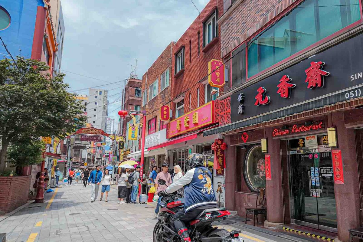 Pedestrians walk along a narrow Chinatown street near Busan Station while a motorcycle passes between restaurants and storefronts. The mixed pedestrian and vehicle space shows why some older Busan streets require closer attention when walking with children.