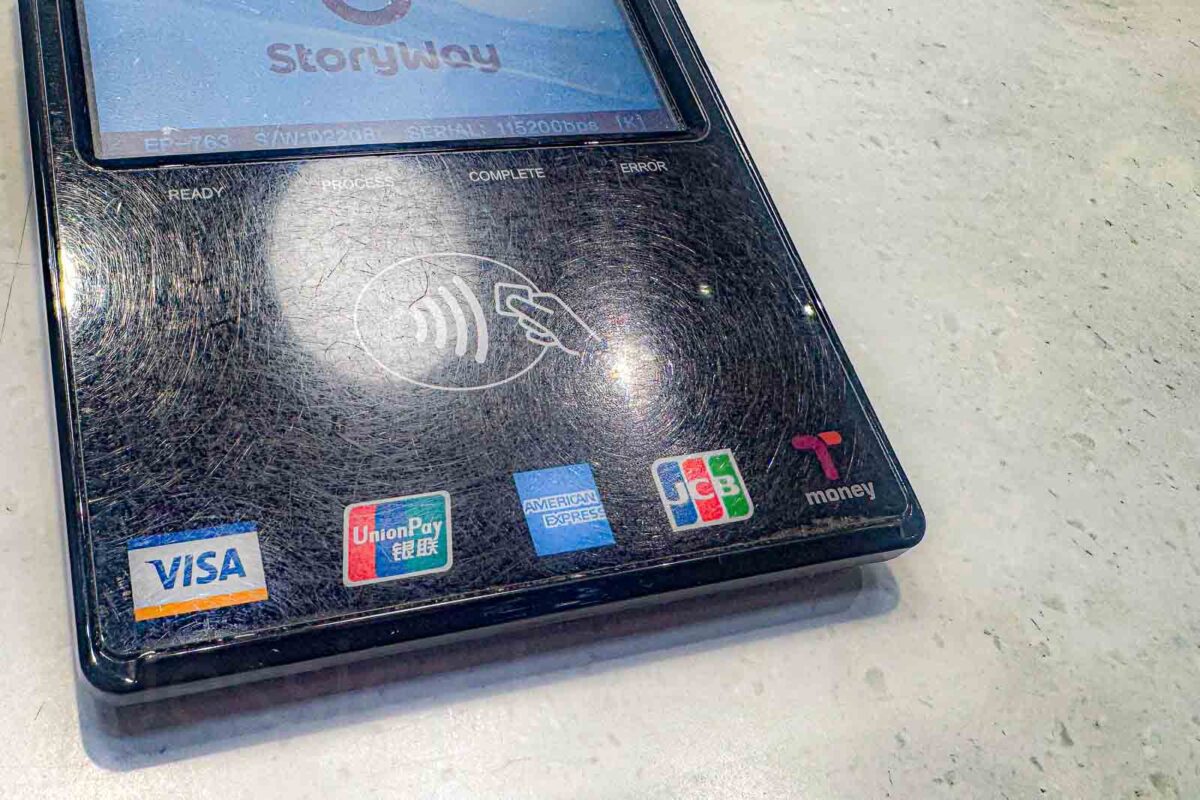 A close view of a StoryWay payment terminal shows contactless payment logos including "VISA". "UnionPay". "AMERICAN EXPRESS". "JCB". "Tmoney". The terminal demonstrates that T-money can also be accepted for purchases outside Busan public transportation.