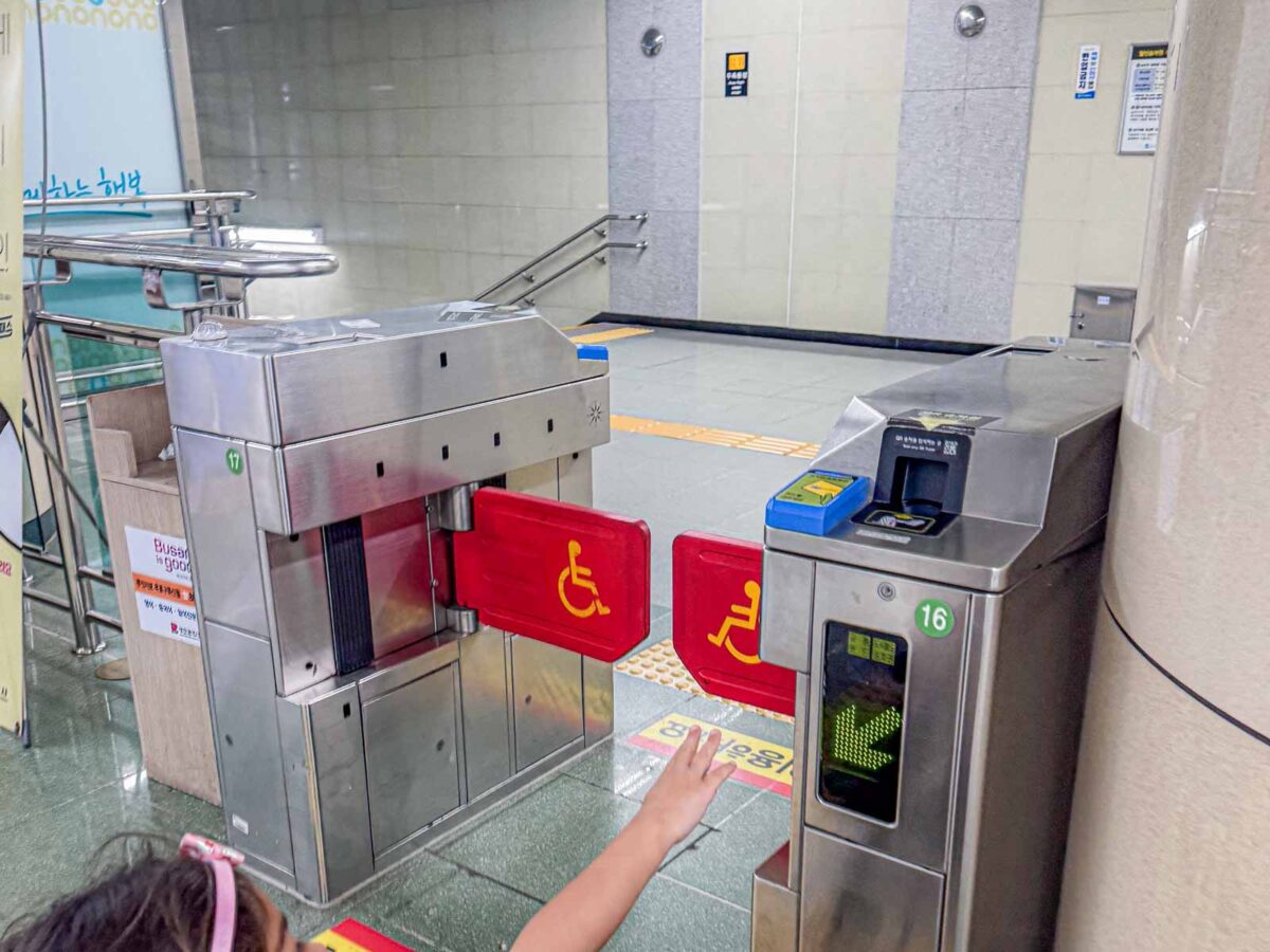 A wide Busan Metro fare gate has red swinging panels marked with a wheelchair symbol. The extra space gives wheelchairs, strollers, and luggage an easier route through the station.