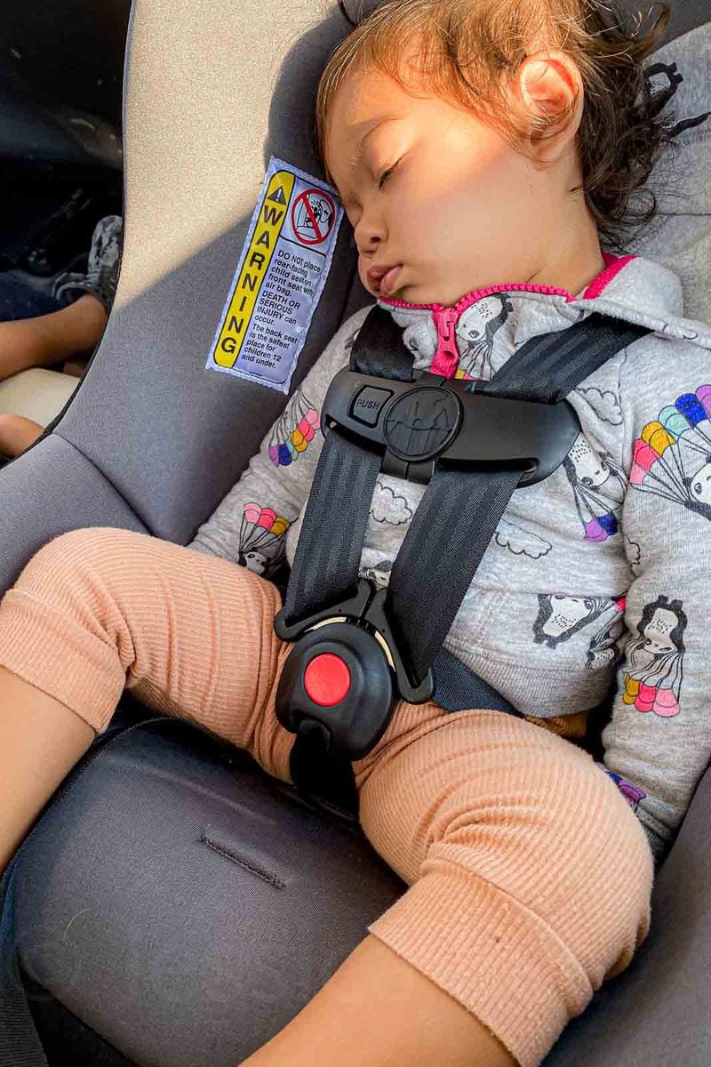 A sleeping toddler rests against the padded side of a gray Cosco travel car seat while secured in the harness. The photo shows the extra structure and head support discussed in the WAYB Pico alternatives section. A yellow safety label beside her head begins with "WARNING".
