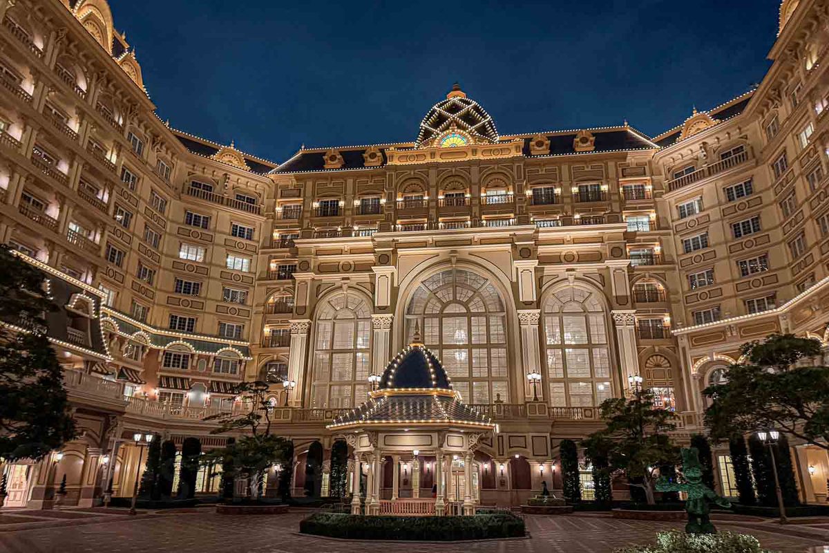 A grand multi-story hotel glows with warm exterior lights around a courtyard under a dark blue night sky. The large property illustrates the range of hotels families may need to investigate when looking for rooms that officially accommodate five people.