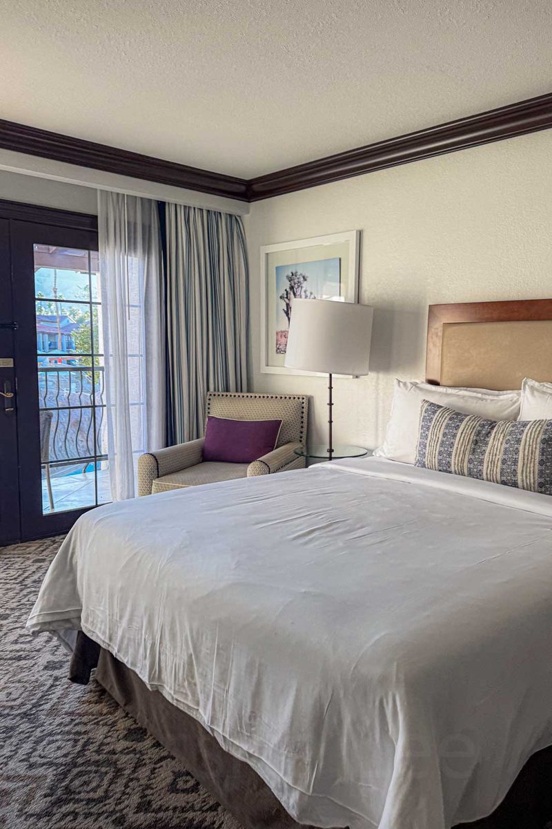 A large white bed fills an Omni Rancho Las Palmas hotel room with an armchair and glass door leading to a balcony. The photo shows why families should study the actual bed layout before deciding whether one hotel room has enough sleeping space.
