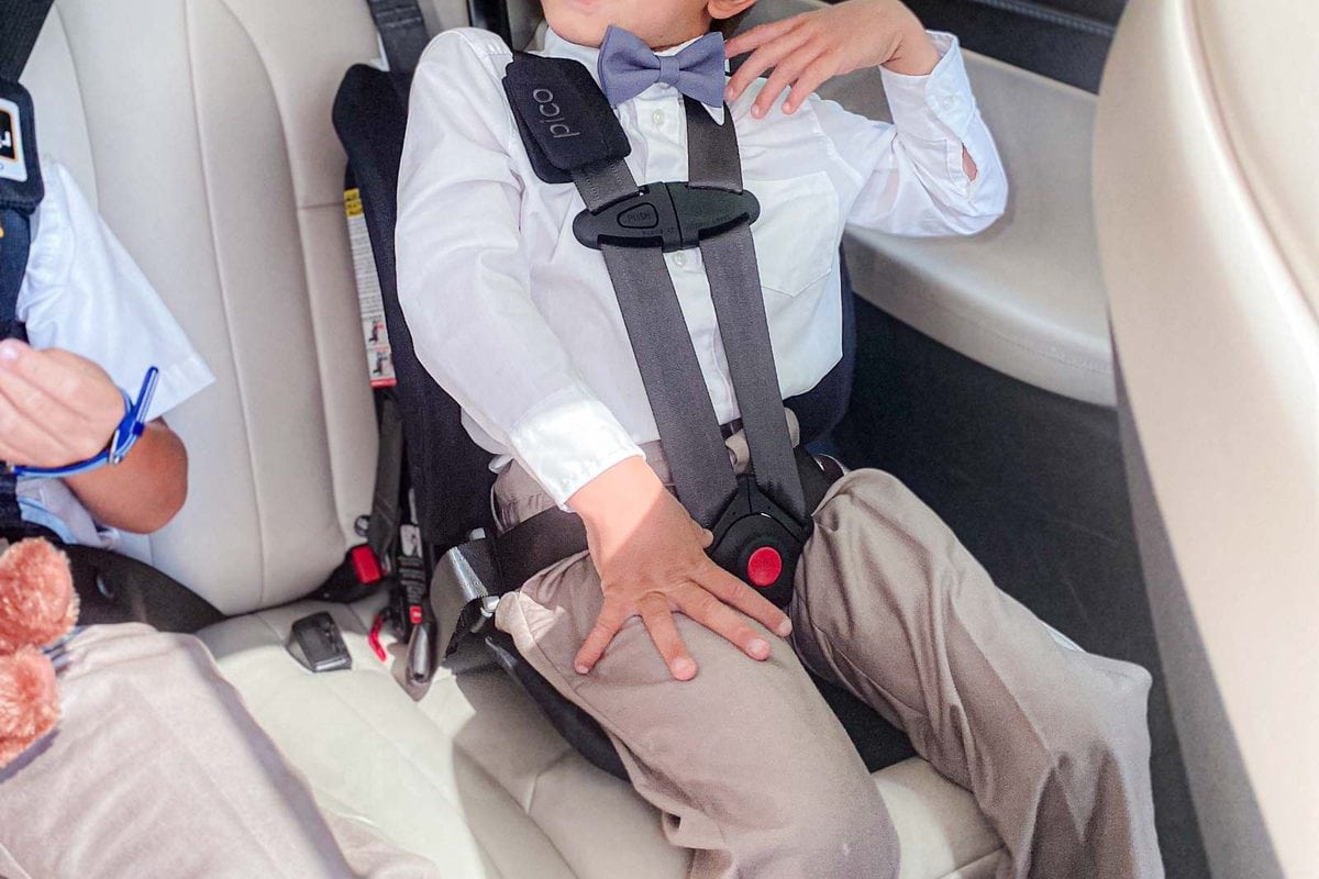 A child wearing a white shirt and bow tie sits buckled into a black Pico car seat on a light colored vehicle seat. The compact design leaves open space beside him and shows the low profile fit of the seat.