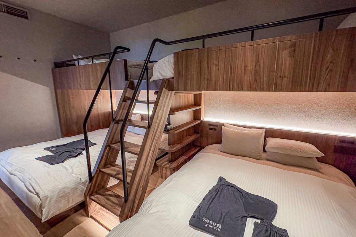 A family room at seven x seven Ishigaki has two large lower beds with two raised sleeping spaces above them and wooden stairs between the beds. The compact layout provides multiple sleeping surfaces in one room for a family of five. Clothing on the beds reads "seven x seven."