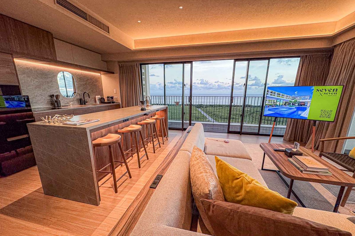 An open plan seven x seven Ishigaki apartment has a full kitchen island, sofa, television, and wide windows overlooking the water. The living space shows how apartment style accommodations can add a kitchen and room to spread out for a family of five. The television displays "seven x seven."