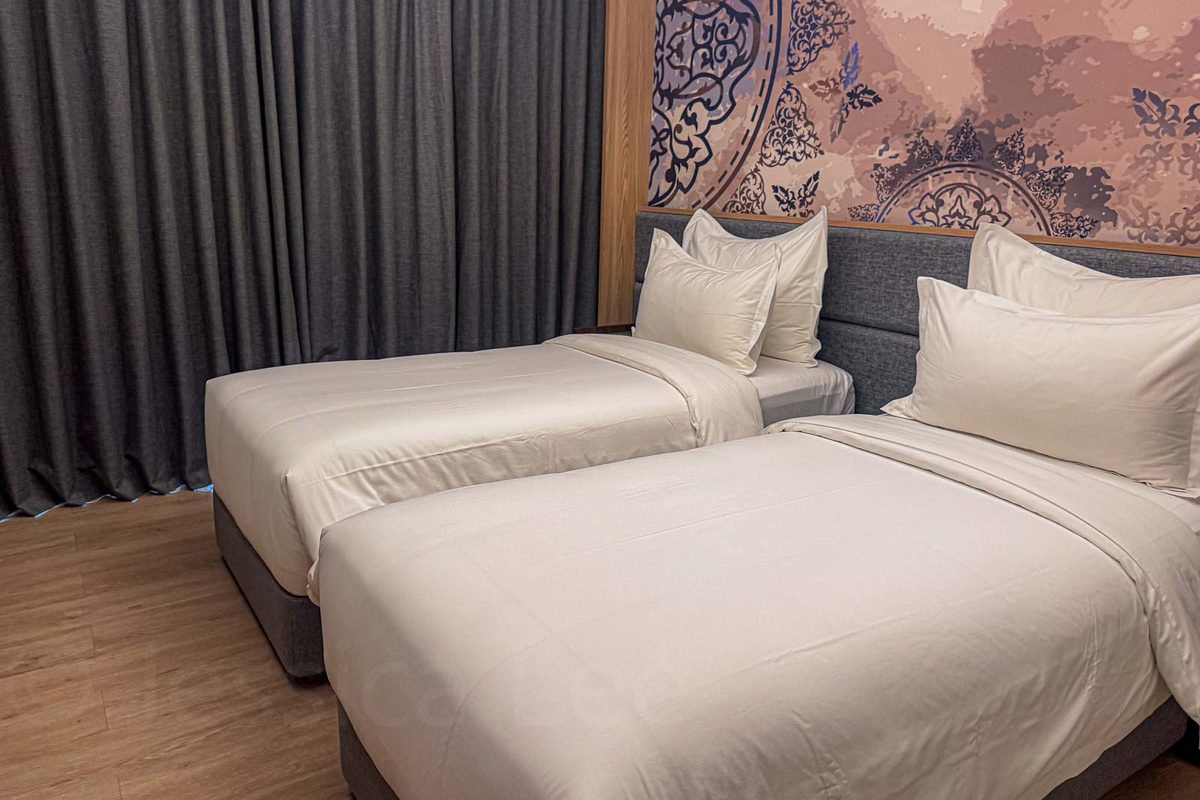Two single beds with white linens sit side by side beneath a patterned wall panel in a hotel room. Separate beds can make a family room more practical when older children no longer want to share one sleeping surface.
