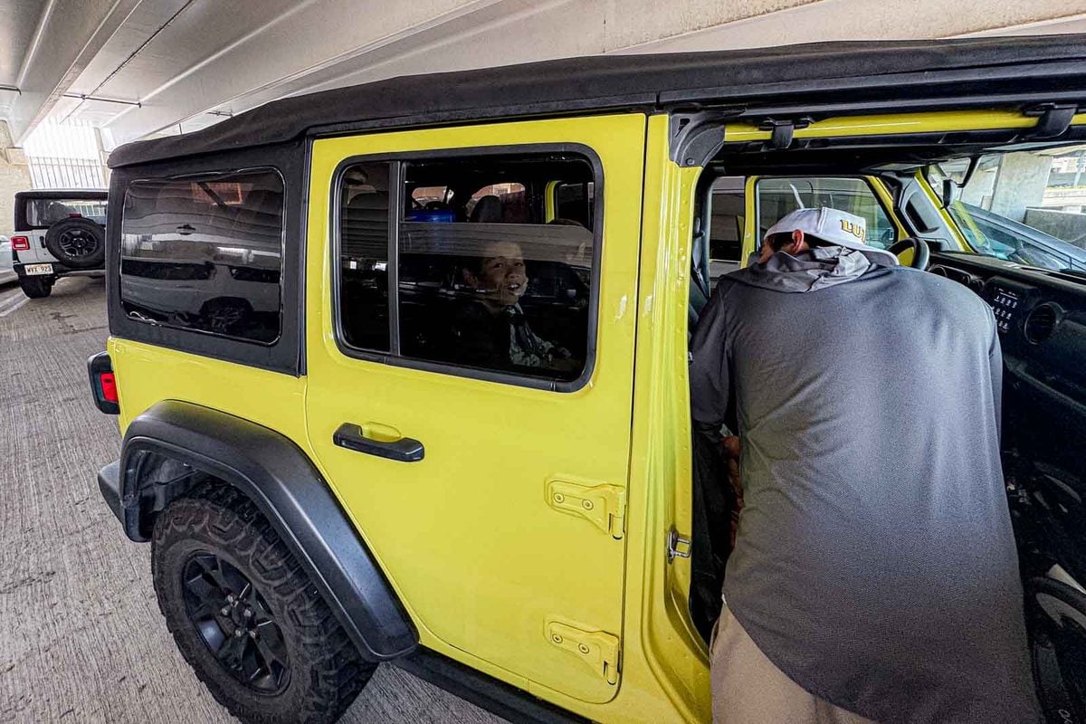 A dad leans into a bright yellow Jeep rental car while a child smiles from the back seat in a parking garage. The rental car scene shows the kind of destination travel where a compact car seat can be easier to manage.