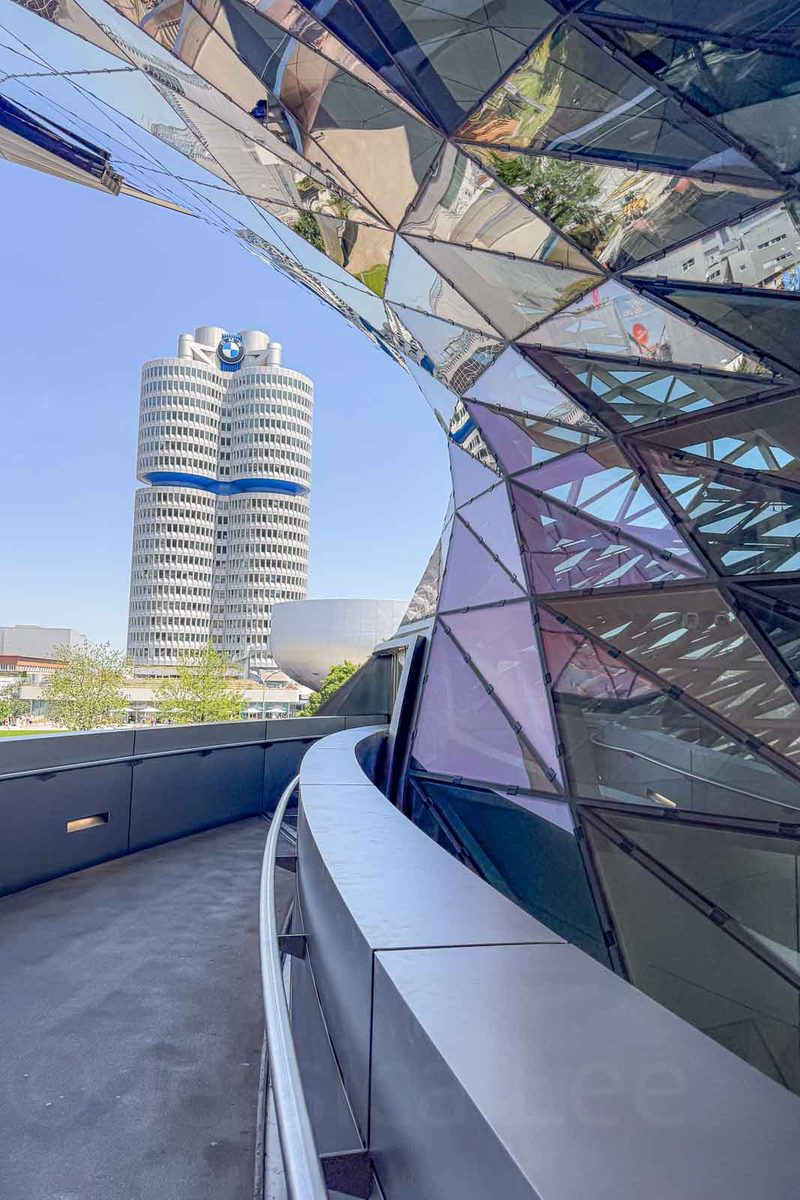 A curved pedestrian walkway alongside the angular glass architecture of BMW Welt in Munich. The distinctive four cylinder BMW headquarters tower stands in the distance against a clear blue sky.