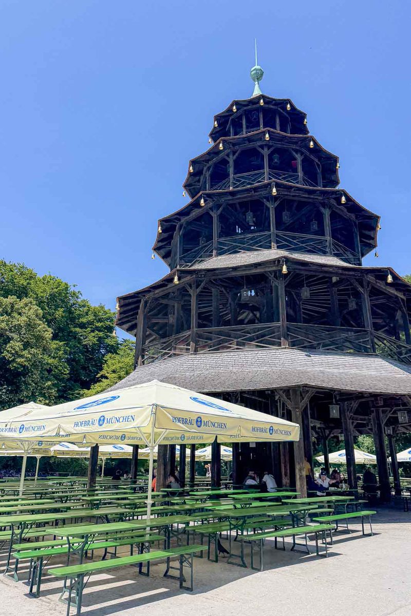 The tall wooden Chinese Tower rises above rows of green picnic tables in the English Garden beer garden in Munich. White umbrellas marked "HOFBRÄU MÜNCHEN" provide shade at this popular outdoor gathering place.