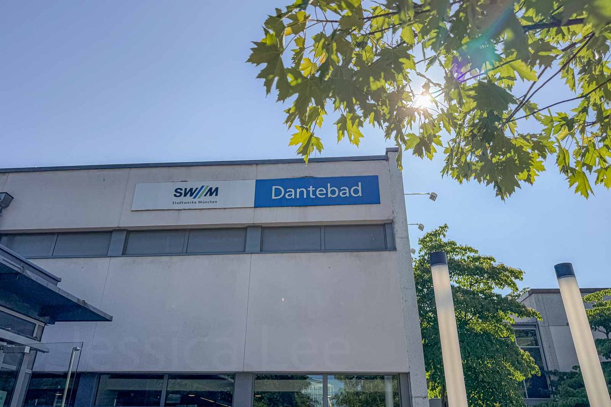 Exterior of the Dantebad public swimming complex in Munich on a sunny summer day. A blue sign reading "Dantebad" sits beside the "SWM" and "Stadtwerke München" logos above the building entrance.