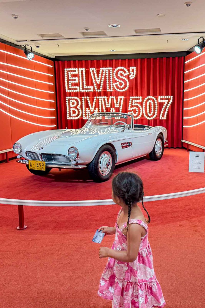 A little girl in a pink floral dress looks at a white vintage BMW convertible displayed on red carpeting at the BMW Museum in Munich. Large illuminated letters behind the car read "ELVIS’ BMW 507".