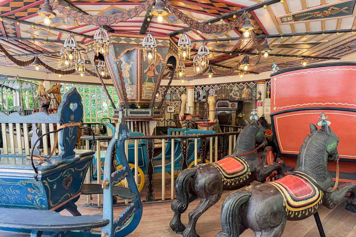 An old fashioned children's carousel with painted wooden horses and colorful carriage seats beneath an elaborately decorated ceiling. This historic carousel near the Chinese Tower playground offers another activity for young kids visiting the English Garden.