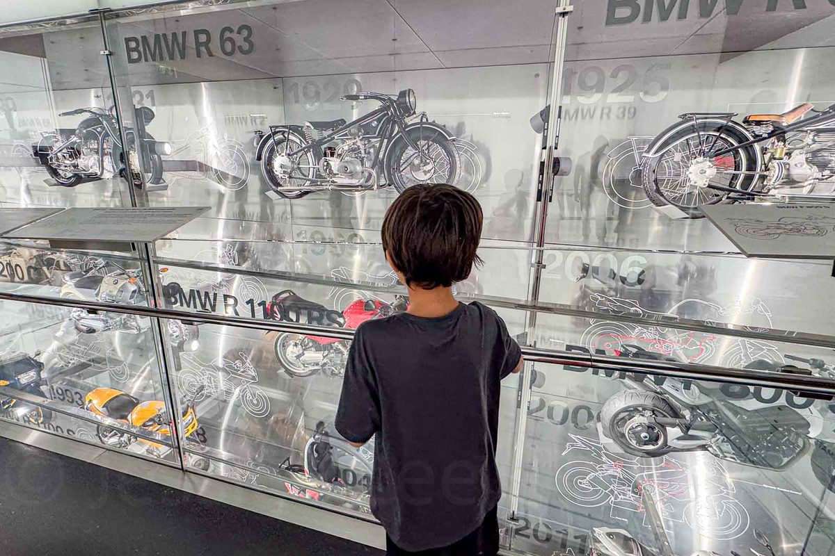 A young boy studies rows of vintage BMW motorcycles displayed behind glass at the BMW Museum in Munich. The exhibit includes historic motorcycle models with labels such as "BMW R 63" and illustrated designs tracing the development of BMW vehicles.