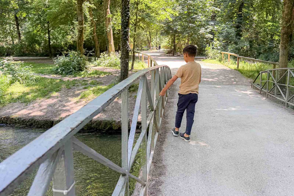 A young boy walks across a small metal bridge over a stream in the English Garden in Munich. Shady walking paths and dense green trees make the park a peaceful place for families to explore.