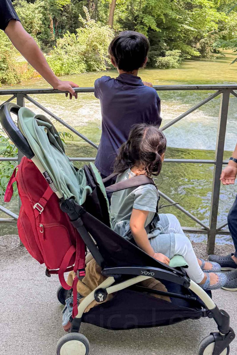 A young girl sits in a compact travel stroller beside an older boy looking over a bridge railing at the flowing water in Munich English Garden. The family pauses along a shaded park path surrounded by greenery.