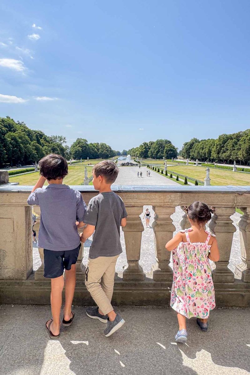 Three children stand at a stone railing overlooking the formal gardens behind Nymphenburg Palace in Munich. A wide pathway stretches toward distant fountains with green lawns and rows of trees on either side.