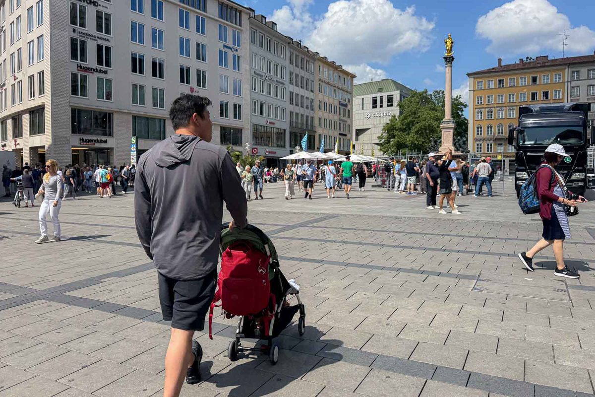 A father pushes a compact travel stroller across the wide pedestrian square of Marienplatz in Munich. Shops and historic buildings surround the plaza with the golden Virgin Mary statue atop a tall column in the background.
