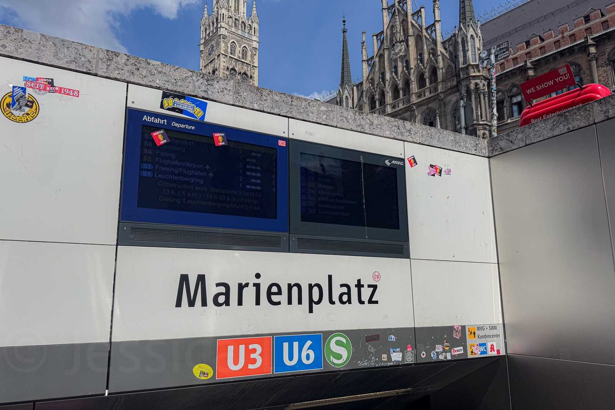 Marienplatz subway station entrance with the Gothic towers of Munich New Town Hall rising behind it. The station sign reads "Marienplatz" with "U3" "U6" and "S" transit symbols beneath departure screens.