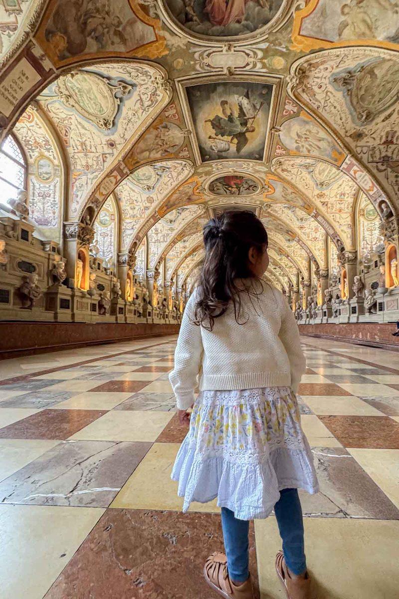 A young girl in a white sweater and patterned skirt stands inside the Antiquarium at Munich Residenz. She looks toward the long hall lined with sculptures and a richly painted vaulted ceiling above a checkered marble floor.