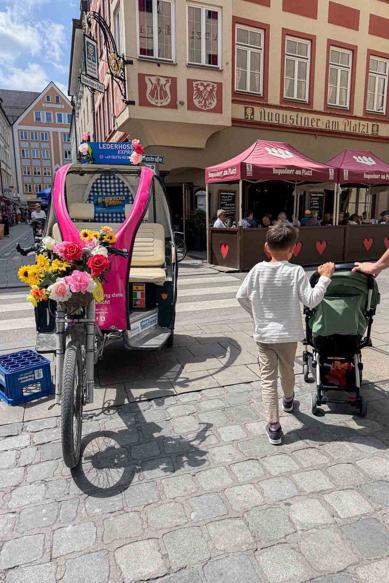 A young boy walks alongside a compact stroller through a cobblestone street in Munich Old Town. A flower decorated bicycle taxi marked "LEDERHOSN EXPRESS" sits beside outdoor restaurant seating at "Augustiner am Platzl".