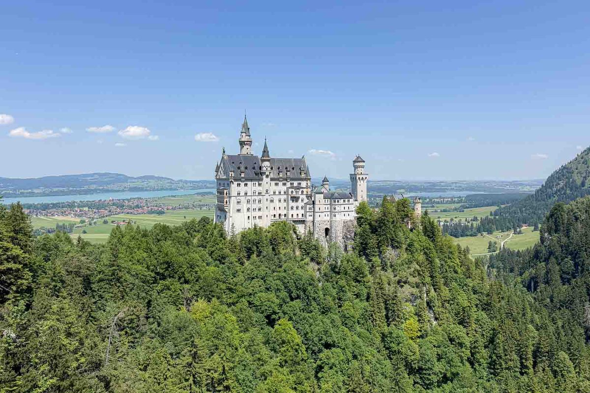 Neuschwanstein Castle rises above a densely forested hillside in southern Bavaria with green valleys and distant countryside behind it. The view from Marienbrücke shows the fairytale castle surrounded by summer scenery.