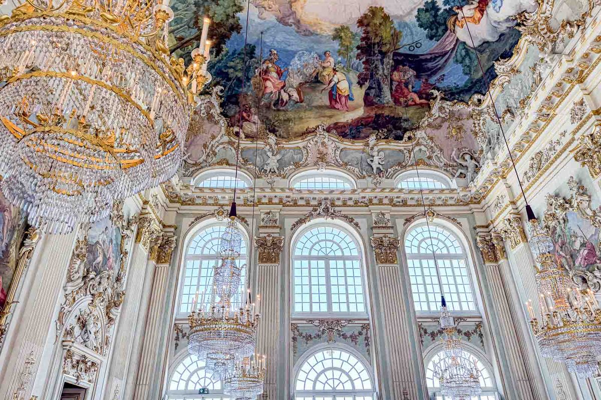 The grand ballroom inside Nymphenburg Palace features massive crystal chandeliers and elaborate gilded decorations. Colorful ceiling frescoes and towering arched windows fill the historic royal room.