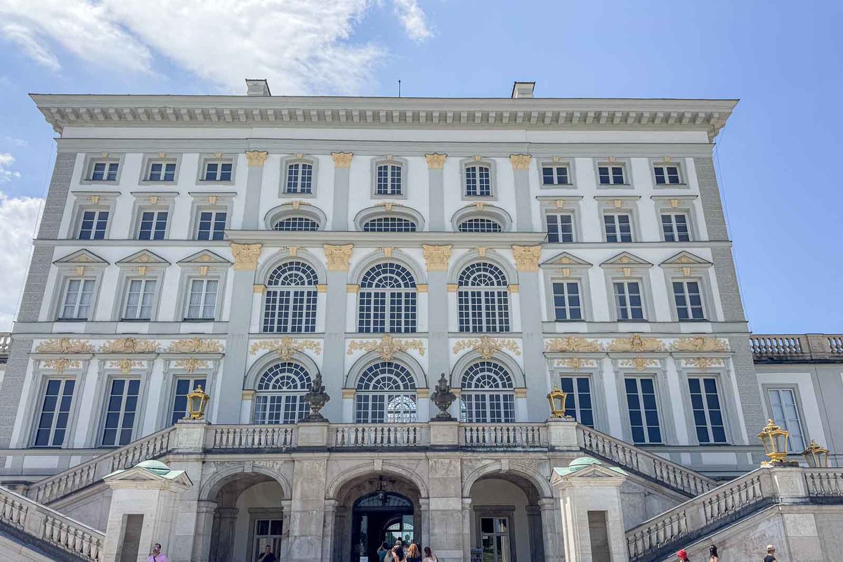 Rear facade of Nymphenburg Palace in Munich with tall arched windows and decorative gold details on the white exterior. Broad stone staircases lead to the palace entrance and surrounding gardens.