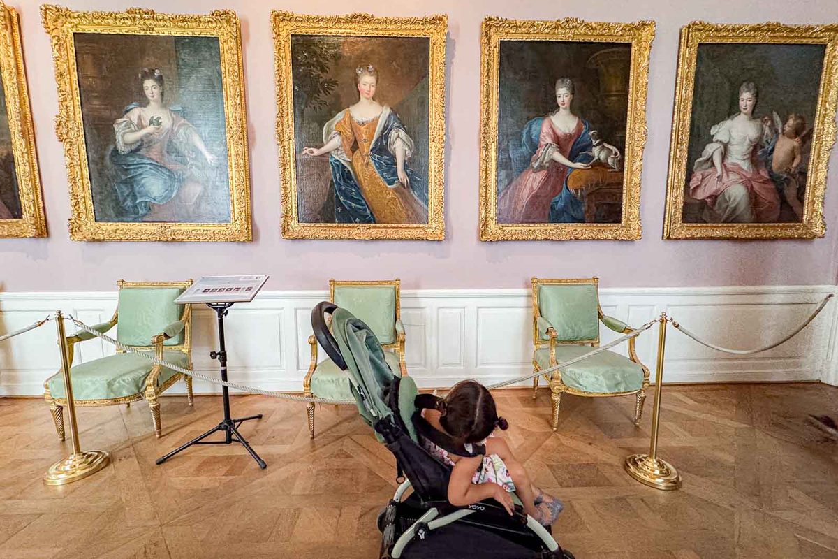 A young girl sits in a lightweight travel stroller inside Nymphenburg Palace in Munich. Four large portraits of royal women hang in ornate gold frames above a row of pale green chairs.
