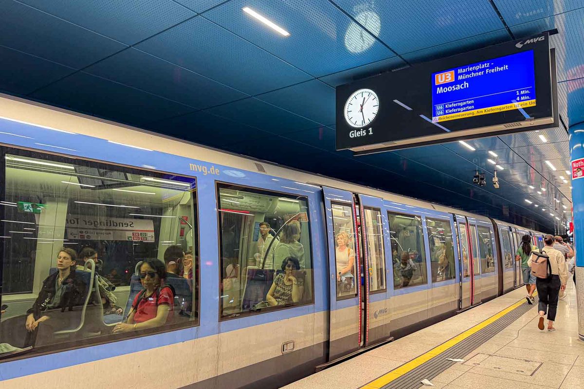 A blue and silver U Bahn train stopped at Rotkreuzplatz station in Munich with passengers visible through the windows. An overhead electronic display shows directions including "Moosach" and "Marienplatz" with estimated train arrival times.