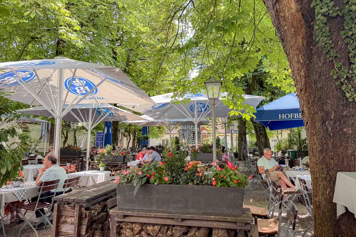 Outdoor dining patio at Schlosswirtschaft Schwaige Nymphenburg in Munich with tables under large white and blue umbrellas. Leafy trees and colorful flower planters create a shaded garden setting for family meals near Nymphenburg Palace.