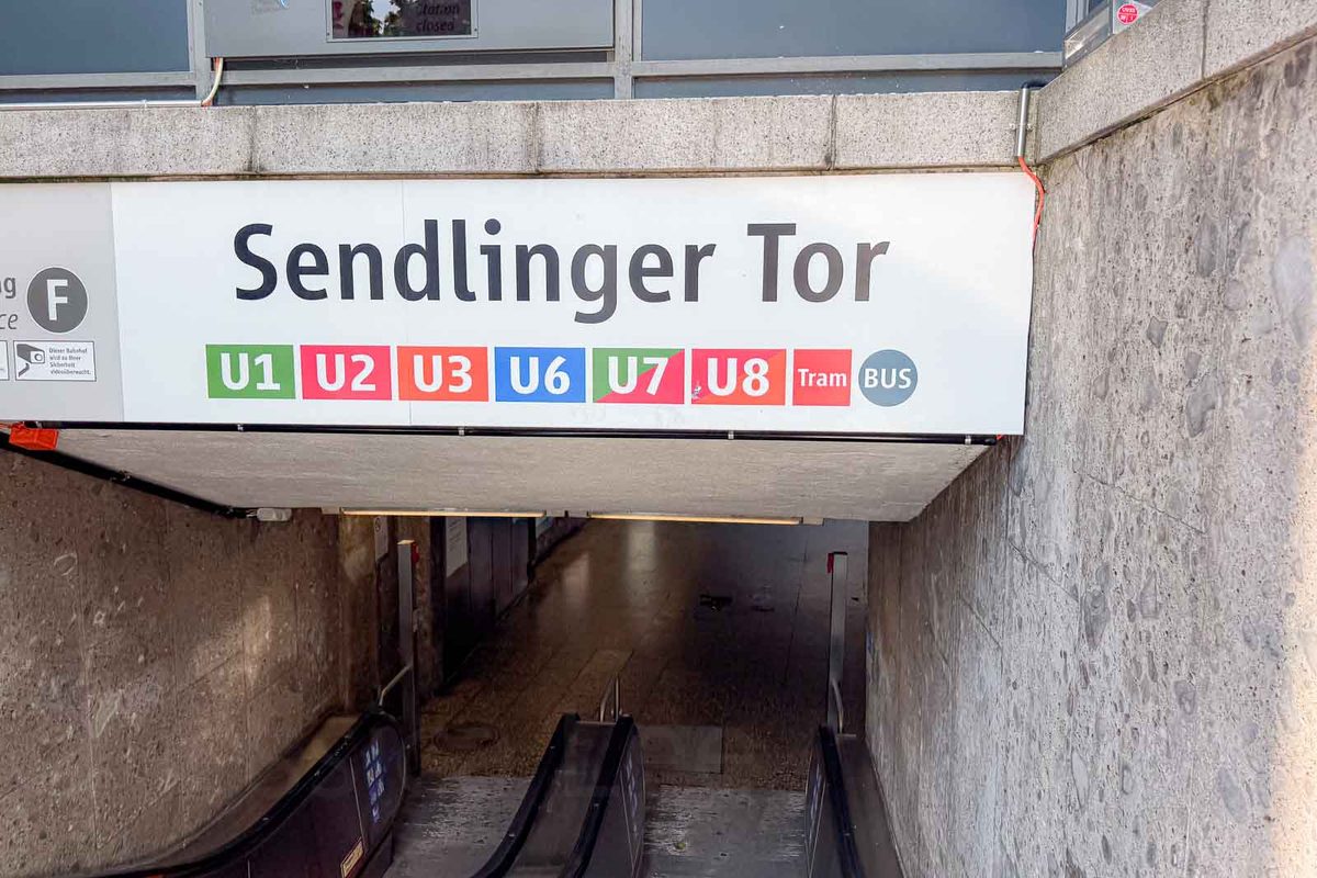 Entrance to Sendlinger Tor subway station in Munich with escalators leading underground. A large transit sign reads "Sendlinger Tor" and shows connections for "U1" "U2" "U3" "U6" "U7" "U8" "Tram" and "BUS".