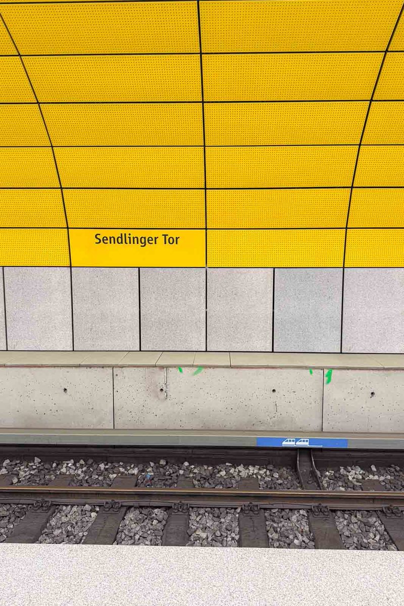 Bright yellow tiled wall at Sendlinger Tor subway station in Munich with the station name "Sendlinger Tor" printed in black. Train tracks run along the platform below the colorful station wall.