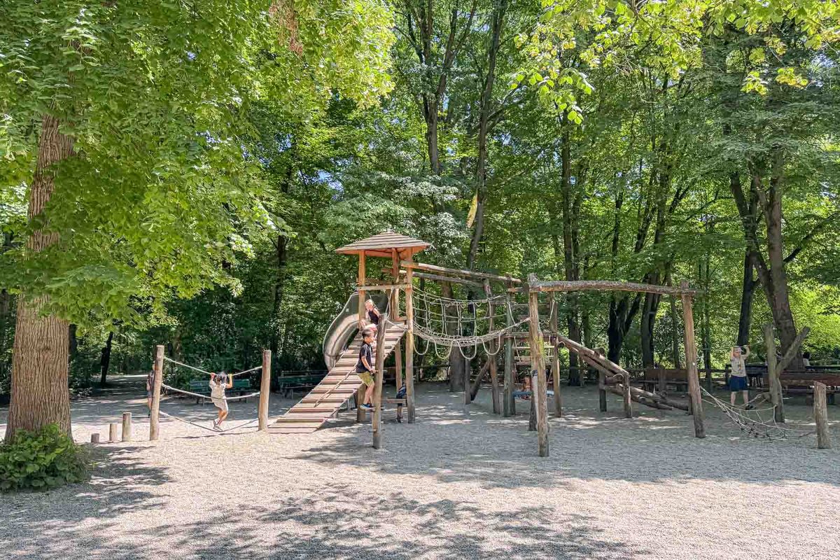 Children play on wooden climbing structures with rope bridges and a metal slide at Spielplatz am Chinesischen Turm in Munich. The large shaded playground sits among leafy trees next to the Chinese Tower beer garden.